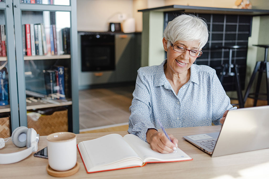 Older woman sitting at computer taking notes in a notebook